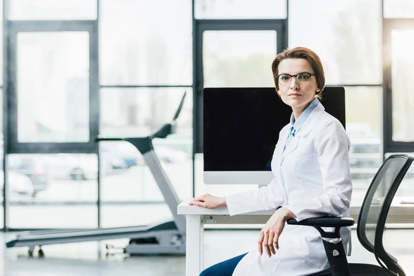 Médico de bata blanca sentado en el escritorio de la computadora en el gimnasio y mirando a la cámara - foto de stock