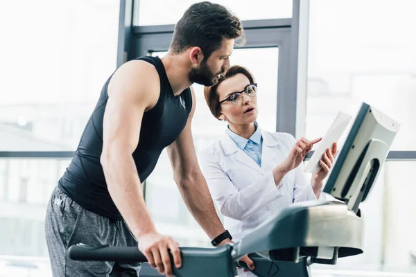 Beau sportif en cours d'exécution sur tapis roulant près du médecin avec tablette numérique pendant le test d'endurance dans la salle de gym — Photo de stock