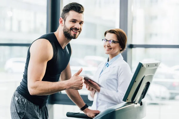 Sportsman running on treadmill and doing thumb up sign near doctor during endurance test in gym — Stock Photo