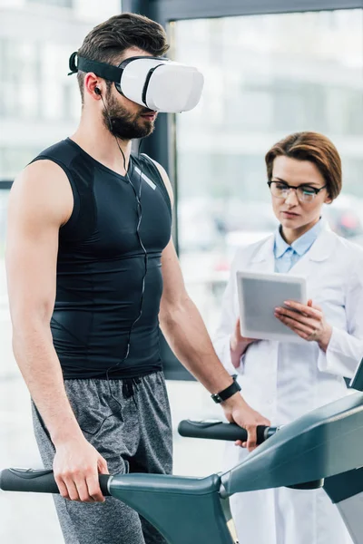 Desportista em vr fone de ouvido correndo em esteira perto do médico durante teste de resistência no ginásio — Stock Photo