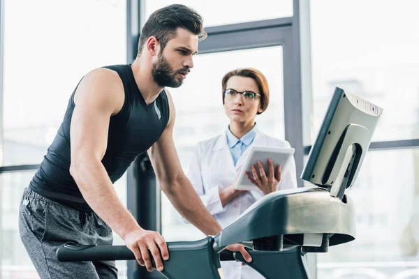 Beau sportif en cours d'exécution sur tapis roulant près du médecin pendant le test d'endurance dans la salle de gym — Stock Photo