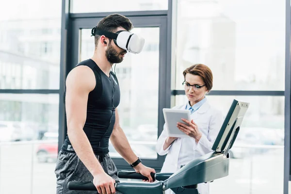 Sportivo in auricolare vr in esecuzione sul tapis roulant vicino al medico durante il test di resistenza in palestra — Foto stock