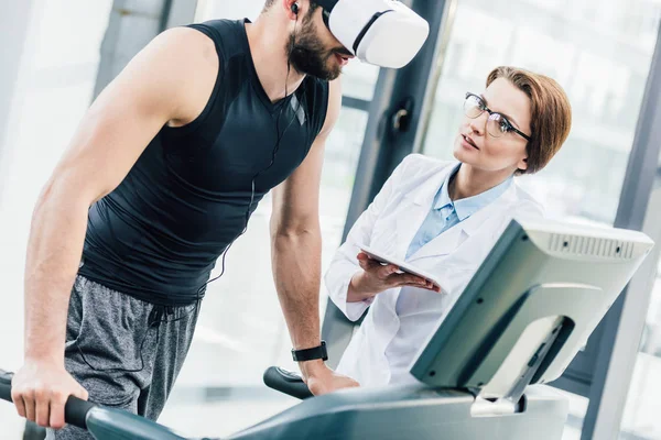 Deportista en entrenamiento de auriculares vr en cinta de correr cerca del médico durante la prueba de resistencia en el gimnasio - foto de stock