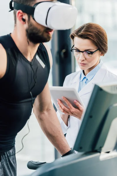 Sportsman running on treadmill near attractive doctor during endurance test in gym — Stock Photo