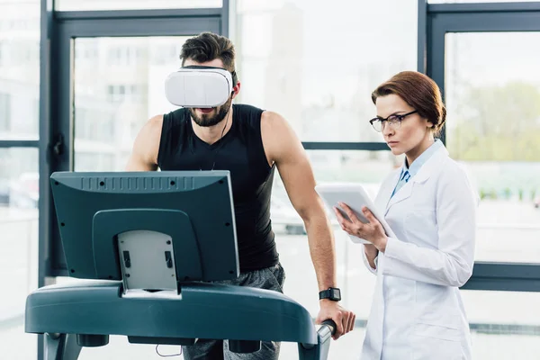 Спортсмен в наушниках виртуальной реальности, работающий на беговой дорожке возле врача во время теста на выносливость — стоковое фото