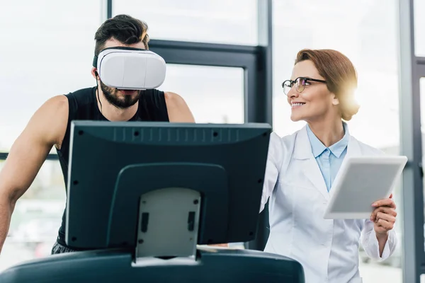 Спортсмен в наушниках виртуальной реальности, работающий на беговой дорожке возле улыбающегося врача во время теста на выносливость — стоковое фото