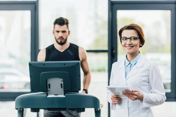 Спортсмен біжить на біговій доріжці біля усміхненого лікаря під час тесту на витривалість у спортзалі — стокове фото