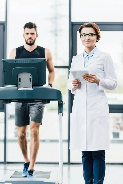 Спортсмен бегает на беговой дорожке рядом с привлекательным врачом во время теста на выносливость в спортзале — стоковое фото