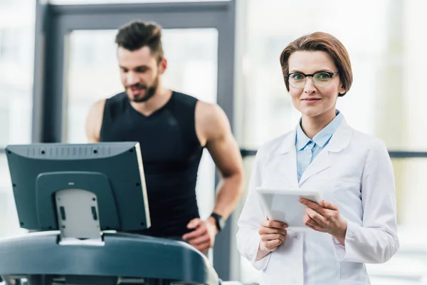 Deportista corriendo en la cinta de correr cerca de atractivo médico durante la prueba de resistencia en el gimnasio - foto de stock