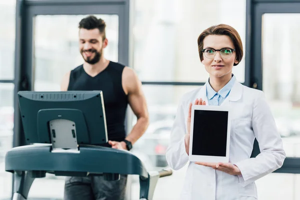 Спортсмен біжить на біговій доріжці біля лікаря, показуючи цифровий планшет під час тесту на витривалість у спортзалі — стокове фото