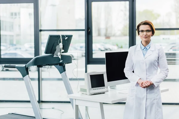 Médecin en manteau blanc près du tapis roulant et bureau d'ordinateur à la salle de gym — Photo de stock