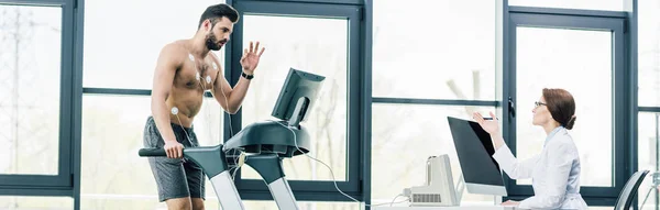 Panoramic shot of sportsman gesturing on treadmill near doctor during endurance test in gym — Stock Photo