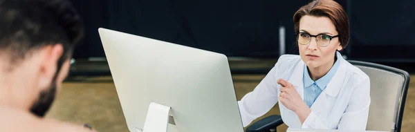 Plano panorámico de hermoso médico sentado en el escritorio de la computadora cerca del paciente - foto de stock