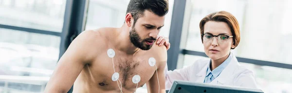 Tiro panorâmico de médico realizando teste de resistência perto de esportista com eletrodos no centro esportivo — Fotografia de Stock