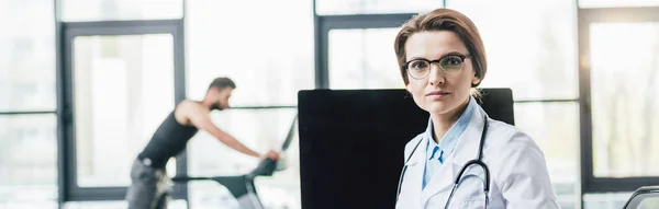 Panoramic shot of beautiful doctor in glasses looking at camera near computer in gym — Stock Photo