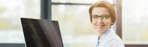 Panoramic shot of beautiful doctor in white coat looking at camera near computer with blank screen — Stock Photo