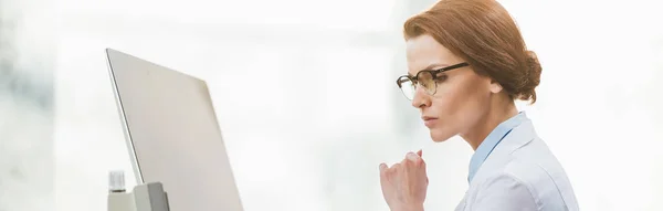 Panoramic shot of beautiful pensive doctor in white coat using computer — Stock Photo