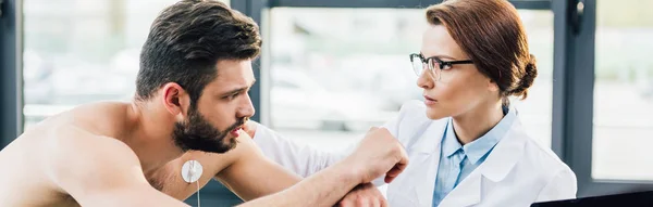 Prise de vue panoramique du médecin effectuant un test d'endurance près du sportif avec des électrodes dans la salle de gym — Photo de stock