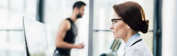 Panoramic shot of beautiful doctor in glasses using computer in gym — Stock Photo