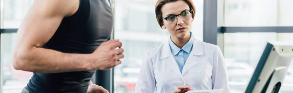 Tiro panorâmico de desportista correndo em esteira perto de belo médico durante teste de resistência no ginásio — Stock Photo