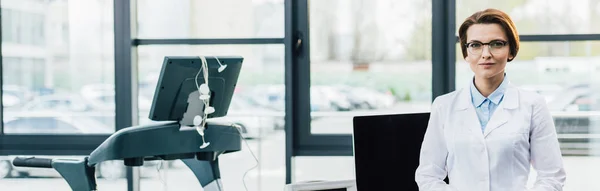 Panoramic shot of doctor in white coat near treadmill at gym — Stock Photo