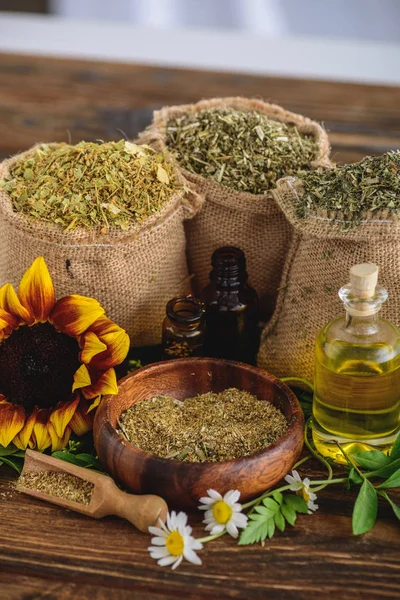 Wooden bowl and sackcloth bags with dried herbs, bottles with essential oils, sunflower and chamomile flowers on wooden surface — Stock Photo
