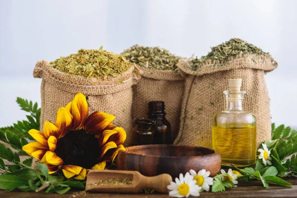 Sackcloth bags with dried herbs, bottles with essential oils, sunflower and chamomile flowers, wooden bowl and spatula on wooden surface — Stock Photo