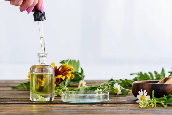 Vista parziale della donna che tiene contagocce vicino bottiglia con olio essenziale, girasole e fiori di camomilla su sfondo bianco — Foto stock