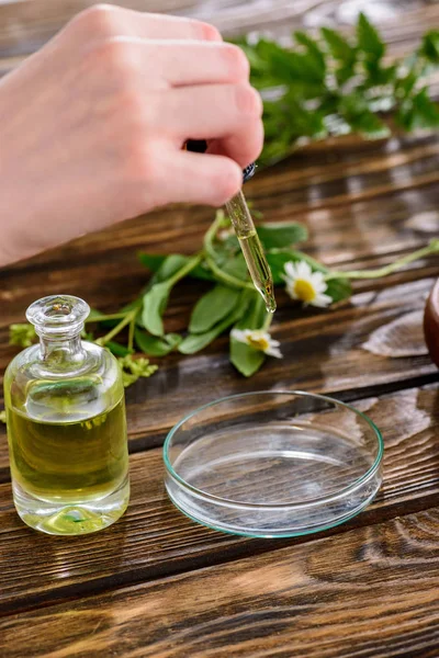 Vista parziale della donna che tiene contagocce vicino alla bottiglia con olio essenziale e ciotola di vetro sulla superficie di legno — Foto stock