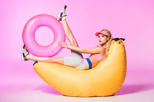 Attractive girl on bean bag chair with inflatable swim ring on pink, doll concept — Stock Photo
