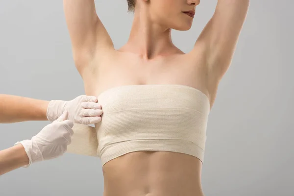 Vista parcial del cirujano plástico en guantes de látex y paciente en vendaje mamario aislado en gris - foto de stock