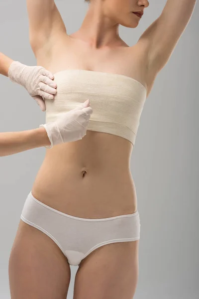 Vue partielle du chirurgien plasticien dans les gants en latex et du patient dans le bandage mammaire isolé sur gris — Photo de stock