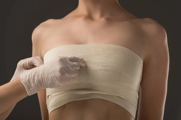 Частичный вид пластического хирурга в латексных перчатках и пациента в грудной повязке, изолированного на сером — стоковое фото