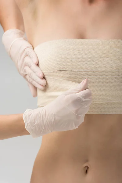 Vista parcial del cirujano plástico en guantes de látex y paciente en vendaje mamario aislado en gris - foto de stock