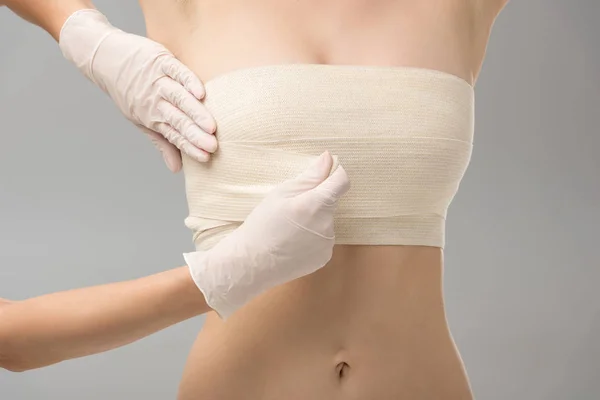 Vue partielle du chirurgien plasticien dans les gants en latex et du patient dans le bandage mammaire isolé sur gris — Photo de stock