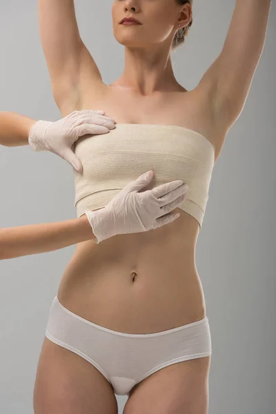 Partial view of plastic surgeon in latex gloves and patient in breast bandage isolated on grey — Stock Photo