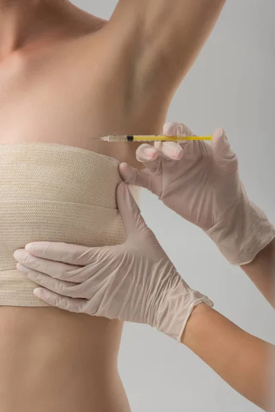 Частичный вид пластического хирурга в латексных перчатках, делающего инъекцию, и пациента в грудной повязке, изолированного на сером — стоковое фото