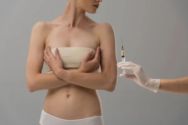 Обрезанный вид пациента с повязкой на груди и врач в латексной перчатке, держащий шприц, изолированный на сером — стоковое фото