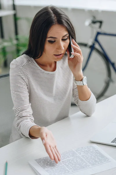 Schöne und brünette Frau, die auf dem Smartphone spricht und auf die Zeitung zeigt — Stockfoto