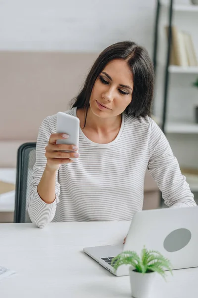Beautiful and brunette woman in jersey using smartphone in apartment — Stock Photo