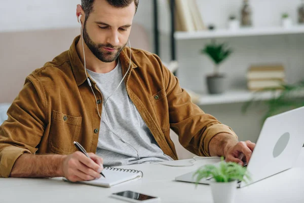 Schöner Mann hört Musik, schreibt in Notizbuch und benutzt Laptop — Stockfoto