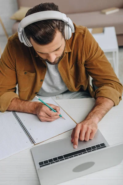 Vista de ángulo alto del hombre escuchando música, escribiendo en el cuaderno y usando el ordenador portátil - foto de stock