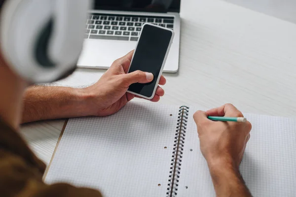 Cropped view of man using smartphone and writing in notebook — Stock Photo