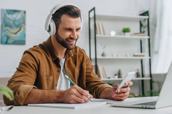 Homem sorrindo usando smartphone e laptop, escrevendo em notebook — Fotografia de Stock