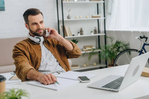 Schöner Mann mit Kopfhörern, der auf den Bildschirm seines Laptops schaut und Bleistift in der Hand hält — Stockfoto