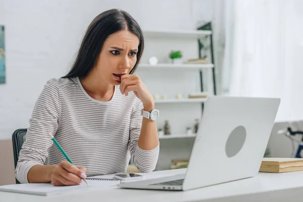 Mujer hermosa y asustada escribiendo en cuaderno con lápiz y mirando a la pantalla de la computadora portátil - foto de stock
