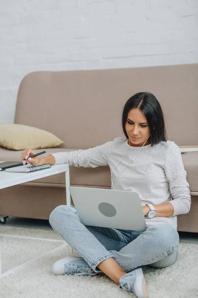Beautiful woman with earphones sitting on floor using laptop and holding pen — Stock Photo