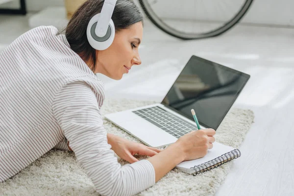 Hermosa mujer con auriculares tumbados en el suelo y la escritura en el cuaderno - foto de stock