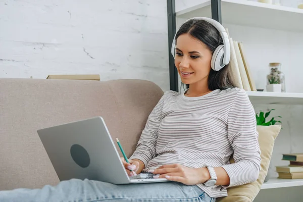 Bella e bruna donna con le cuffie sorridenti e guardando lo schermo del computer portatile — Foto stock