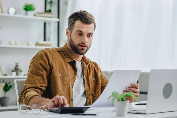 Hombre guapo en camisa sosteniendo papel y calculando con calculadora - foto de stock